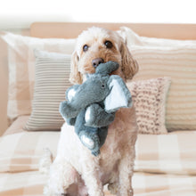 Load image into Gallery viewer, A small cream-colored dog sits on a bed holding a plush elephant toy in its mouth. The background features neutral-toned pillows and a checkered bedspread.