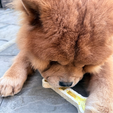 Load image into Gallery viewer, A fluffy brown dog lying on gray stone pavement, chewing on a bone-shaped treat.
