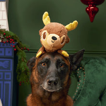 Load image into Gallery viewer, A brown German Shepherd dog sits in a green armchair wearing a small plush reindeer toy on its head, set against a dark green wall with holiday decorations.
