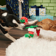 Load image into Gallery viewer, Two dogs tug-of-war with a Christmas-themed rope toy featuring red ornaments and green stocking shapes, set on a white fluffy rug with wrapped gifts and a green brick wall in the background.

