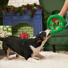 Load image into Gallery viewer, A black and white dog lies on a white shag rug, playfully biting a green Christmas wreath-shaped dog toy held by a person in a holiday-themed room with a fireplace backdrop.
