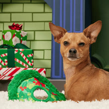Load image into Gallery viewer, A small tan dog sits on a white rug next to a green plush pet hat with a Christmas holly pattern, with wrapped holiday gifts and a green brick-patterned backdrop in the background.
