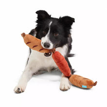 Load image into Gallery viewer, A black and white dog lies on a white studio background holding a plush toy shaped like hot dogs in its mouth.
