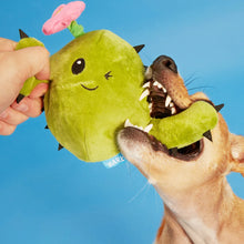 Load image into Gallery viewer, A green plush cactus dog toy with a pink flower on top, being held by a hand and chewed by a dog against a bright blue background.
