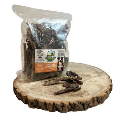 Packaged Camel Tripe dog treats on a wooden surface, with some treats visible outside the package.