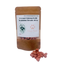 Load image into Gallery viewer, A stand-up kraft paper pouch labeled 'Ocean Omega Krill Training Treats 100g' with a clear window showing reddish-brown dog treats inside, plus a small pile of the same treats in front on a white background.
