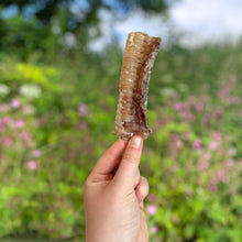 Load image into Gallery viewer, Hand holding a crystal against a blurred natural background with greenery and flowers
