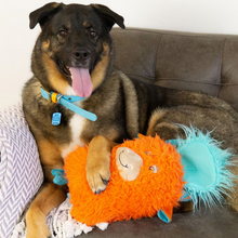 Load image into Gallery viewer, A black and tan dog lying on a brown leather sofa, holding an orange plush monster toy with turquoise accents. The dog is wearing a blue collar, and a light purple patterned throw blanket is draped over the arm of the sofa.
